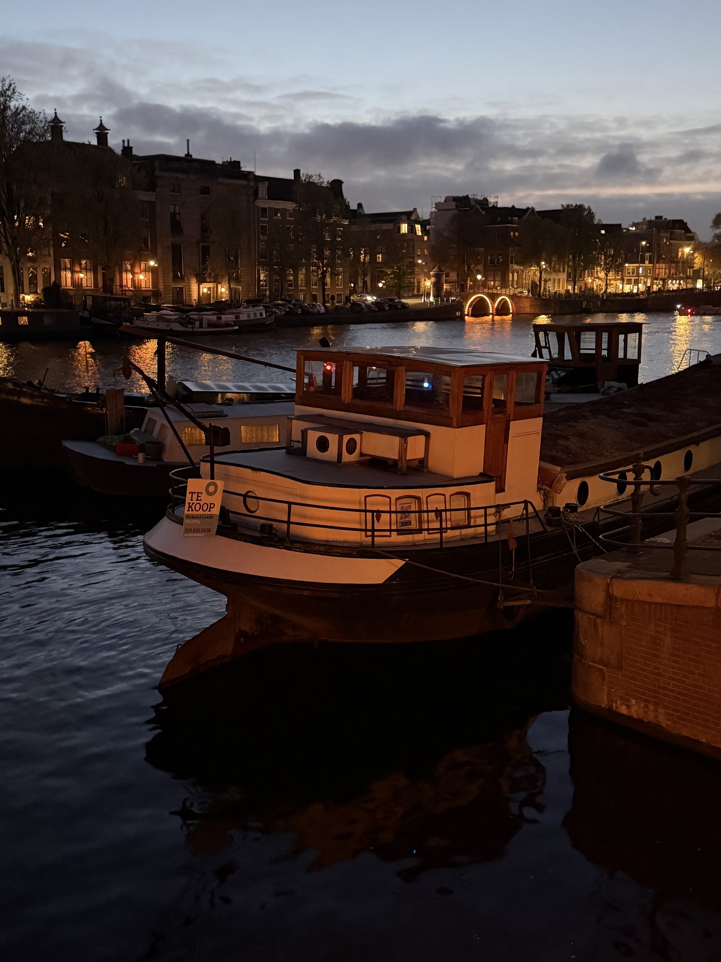 The Amstel river in Amsterdam's Weesperbuurt is captured at dusk, with a prominent white boat displaying a 'TE KOOP' sign
